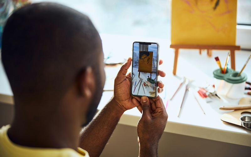 A man with dark skin and a shaved head is seen from behind, holding a smartphone in his hands. He is taking a photo of his artwork, which is displayed on a small easel on a windowsill. The painting features abstract brushstrokes on a yellow canvas. Art supplies, including paintbrushes, pencils, and other tools, are scattered on the table in front of him. Natural light streams in from the window, illuminating the workspace.