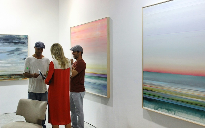 Three people—two men and a woman—stand in conversation inside a contemporary art gallery, surrounded by large abstract landscape paintings with soft, blurred colors.