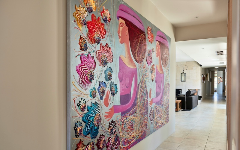 A large, colorful painting of two women surrounded by vibrant floral patterns is displayed in a modern, sunlit hallway with tiled floors and neutral walls.