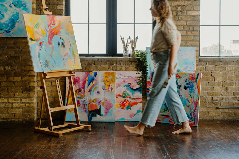 A barefoot artist, Alison Jones Hunt, walks across a well-lit studio space with exposed brick walls and large windows. She is wearing a beige top and light-wash jeans, holding a paintbrush in her hand. A wooden easel holds a colorful abstract painting with fluid brushstrokes in shades of blue, pink, and yellow. Several more vibrant abstract artworks are propped against the wall, adding to the creative atmosphere. Jars filled with paintbrushes sit on the windowsill
