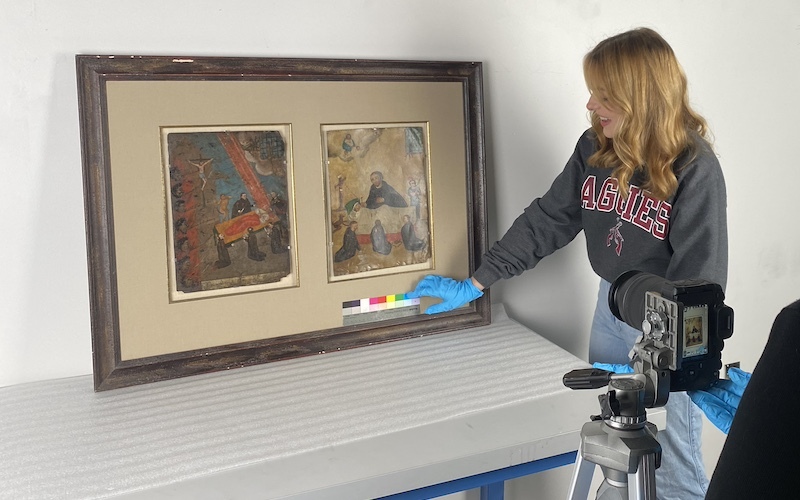 Young woman with latex gloves holds a tool underneath two artworks within a frame; in the corner is a camera capturing the framed work