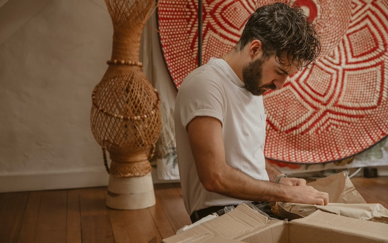 White man with a beard sits on the floor with an open cardboard box with two textile artisan pieces behind him