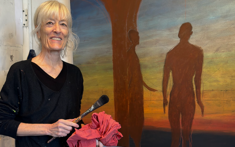 This image features artist Monique Crepault standing in front of one of her paintings. She holds a paintbrush and a red cloth while smiling at the camera. The painting behind her depicts silhouetted human figures in earthy tones against a gradient sky. Monique is dressed in a black, paint-splattered top.