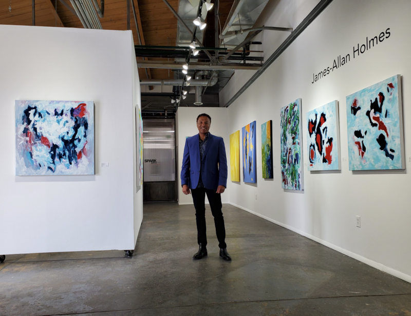 This image shows artist James-Allan Holmes standing in a contemporary art gallery surrounded by his vibrant abstract paintings. The gallery space features clean white walls, high industrial ceilings, and well-placed lighting that highlights the colorful works. Holmes, dressed in a sharp blue blazer and black pants, stands confidently in the center of the room.