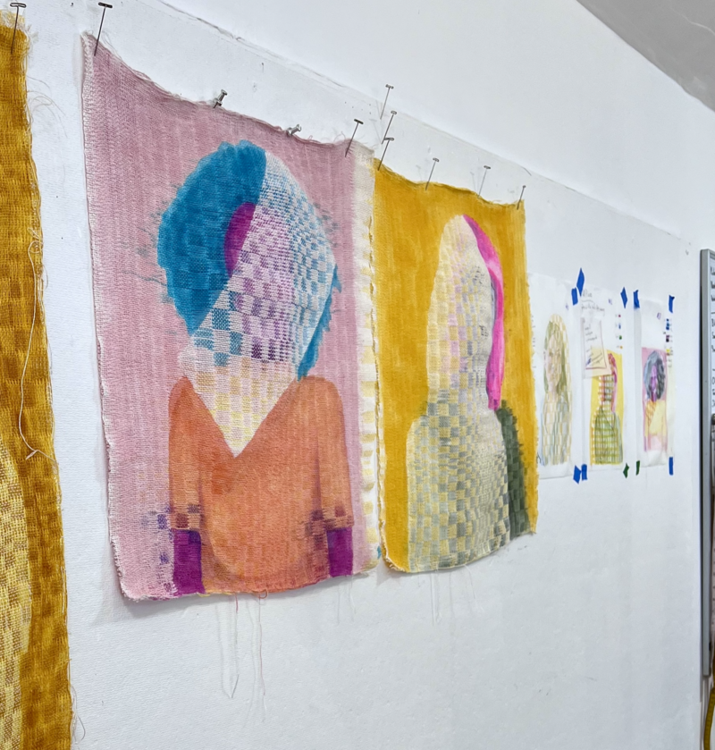 Textile artwork by artist Melissa English Campbell is displayed on a studio wall. The pieces feature abstract, faceless human figures in colorful, checkered patterns. The artwork combines vibrant tones like pink, blue, orange, and yellow, with layered textures resembling woven fabric. The textiles are pinned to the wall, accompanied by smaller sketches or studies taped alongside